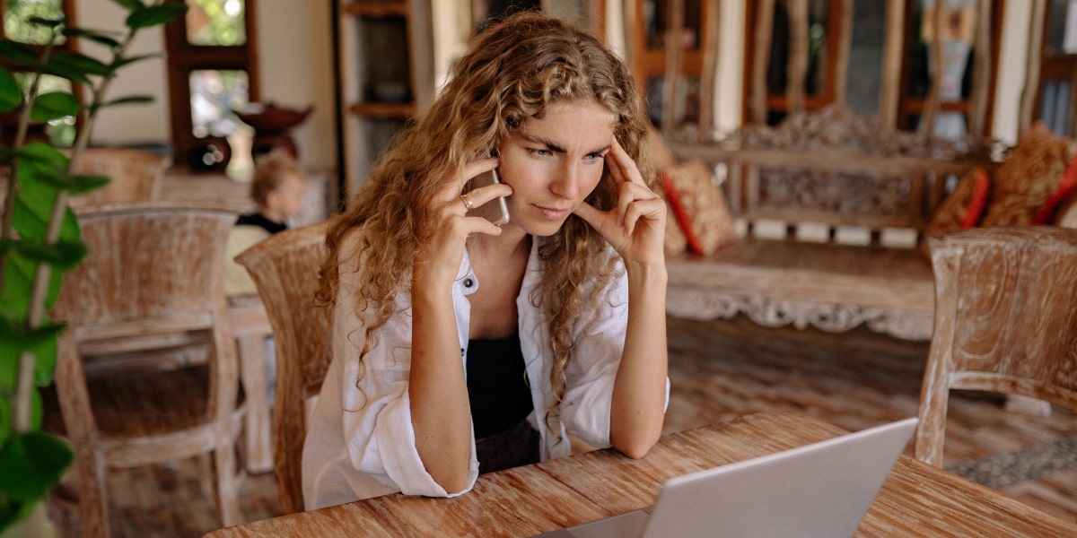 Someone sitting at their dining table, looking at their laptop, and talking on the phone, looking frustrated