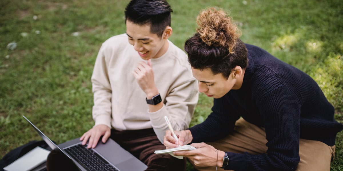 Two designers sitting outside on the grass, working on a laptop and taking notes